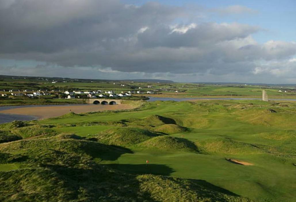 Links greens winding through the dunes above the estuary, with the village and its stone bridge beyond, County Clare